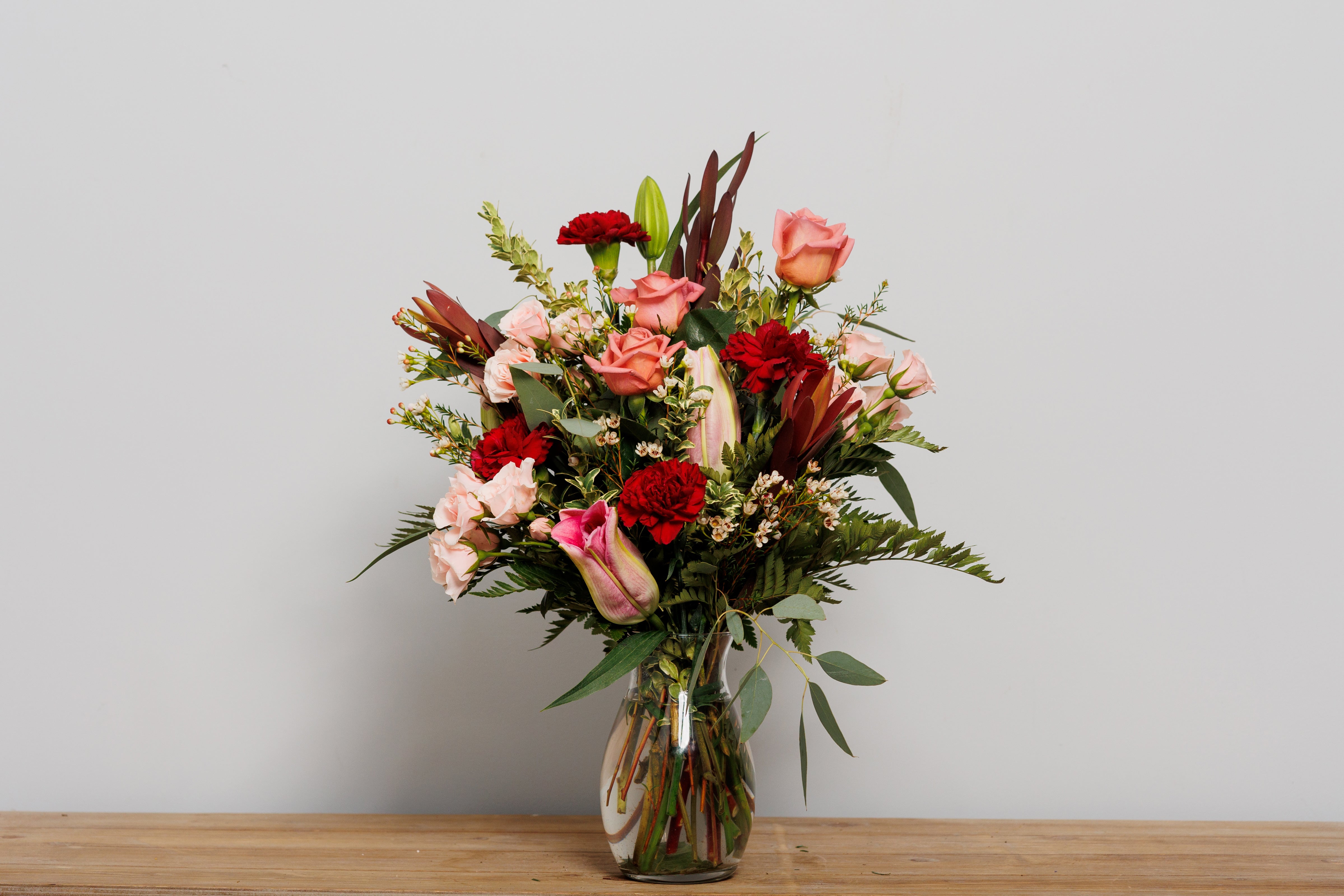 A vase arrangement with dark red carnations, pink lilies and pink roses.