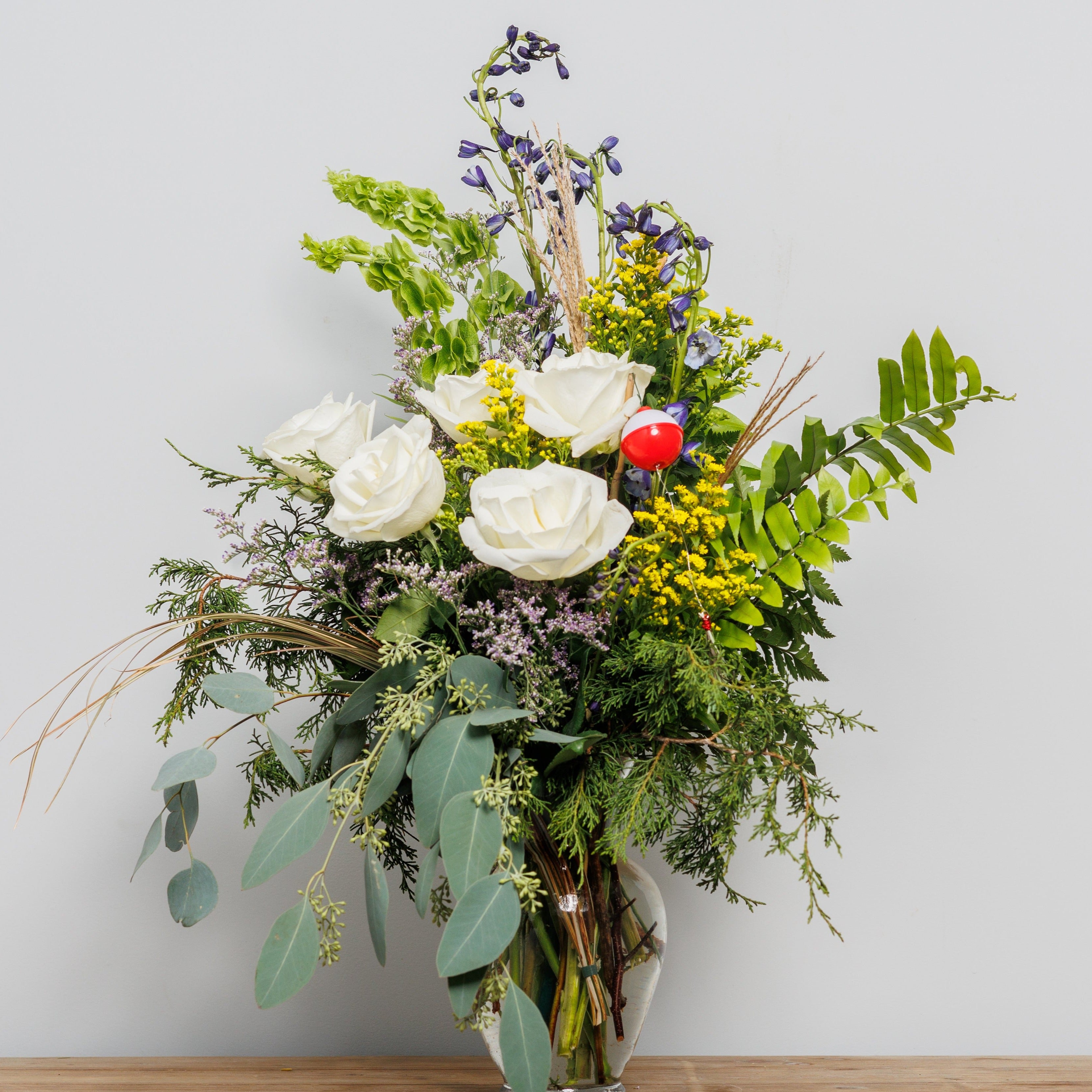A vase arrangement with lots of greenery and white roses with a fishing accessory.