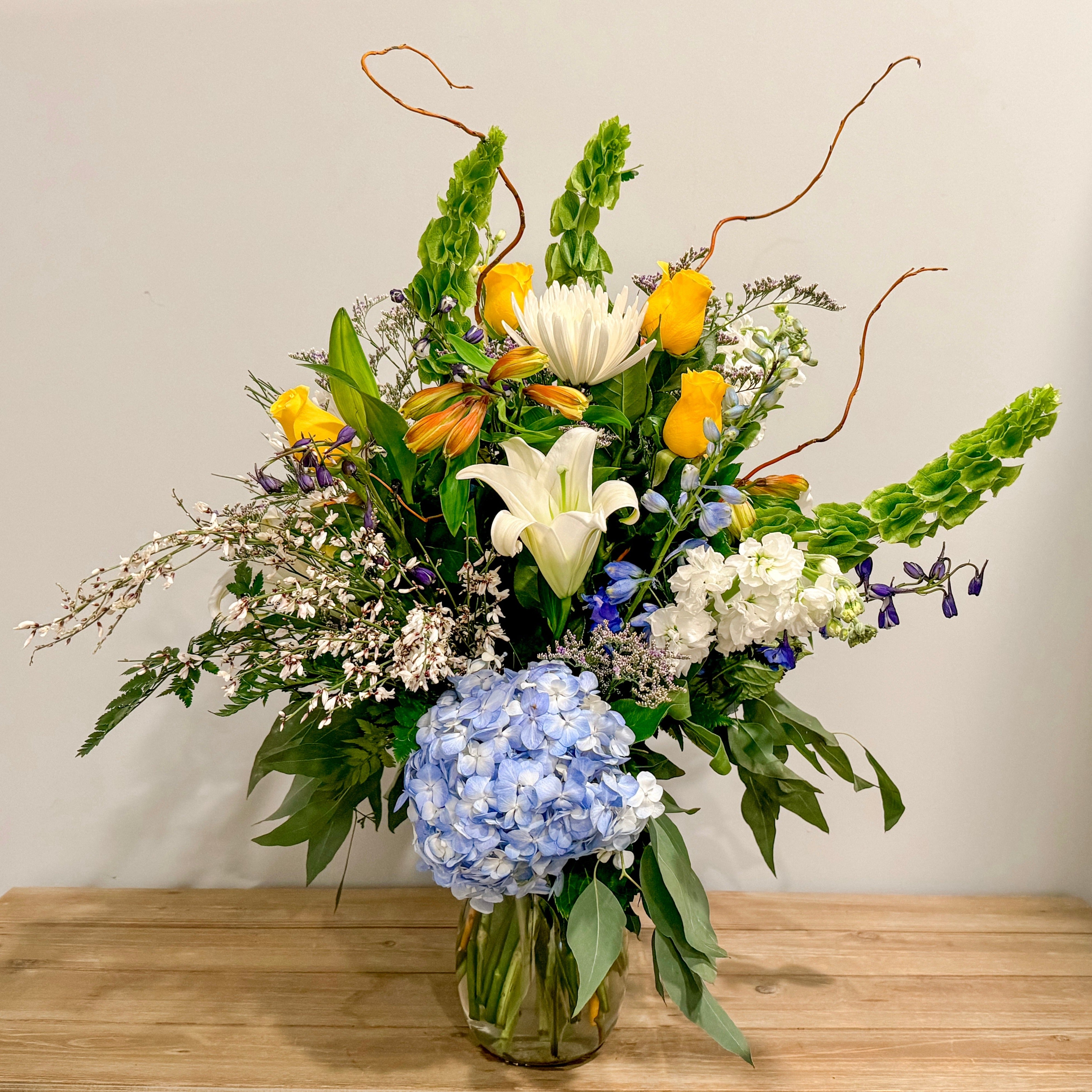 A vase arrangement with yellow roses, bells of Ireland, blue hydrangea and curly willow.