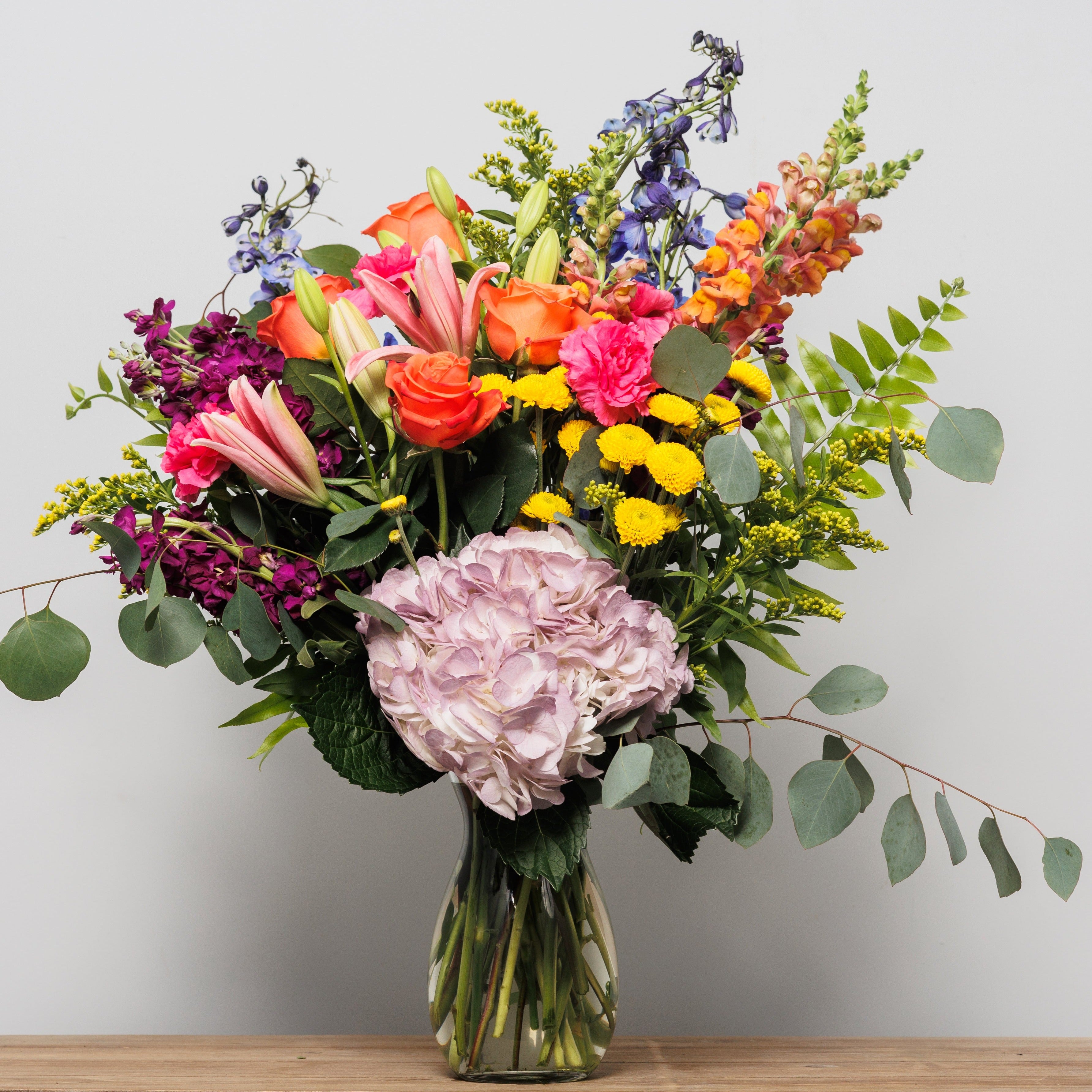 A vase arrangement with orange roses, pink lilies, orange snapdragons, delphinium, stock and hydrangea.