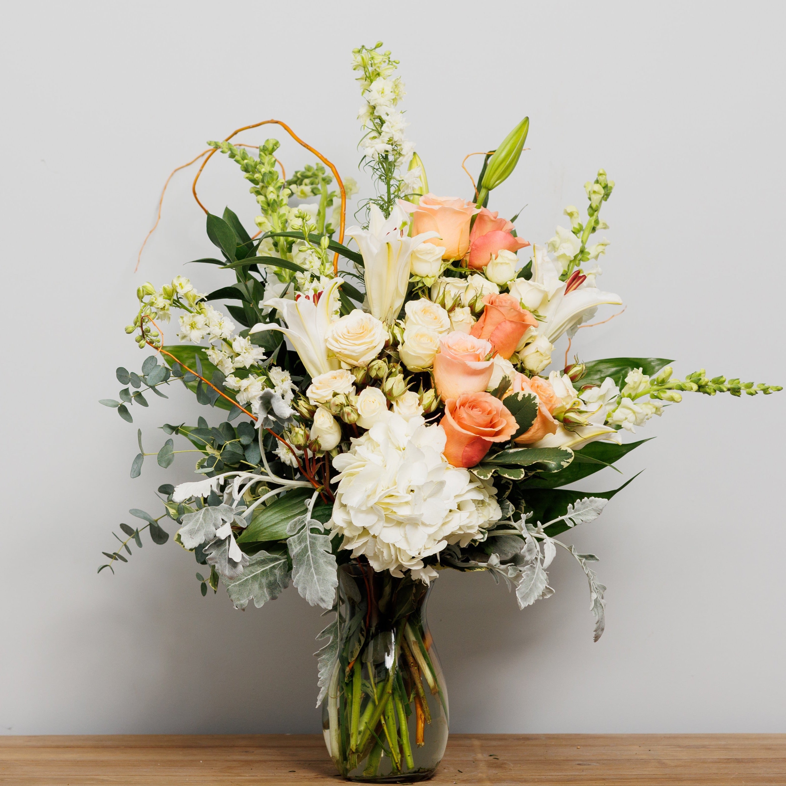 A vase arrangement with a white hydrangea, peach roses, white lilies and curly willow.