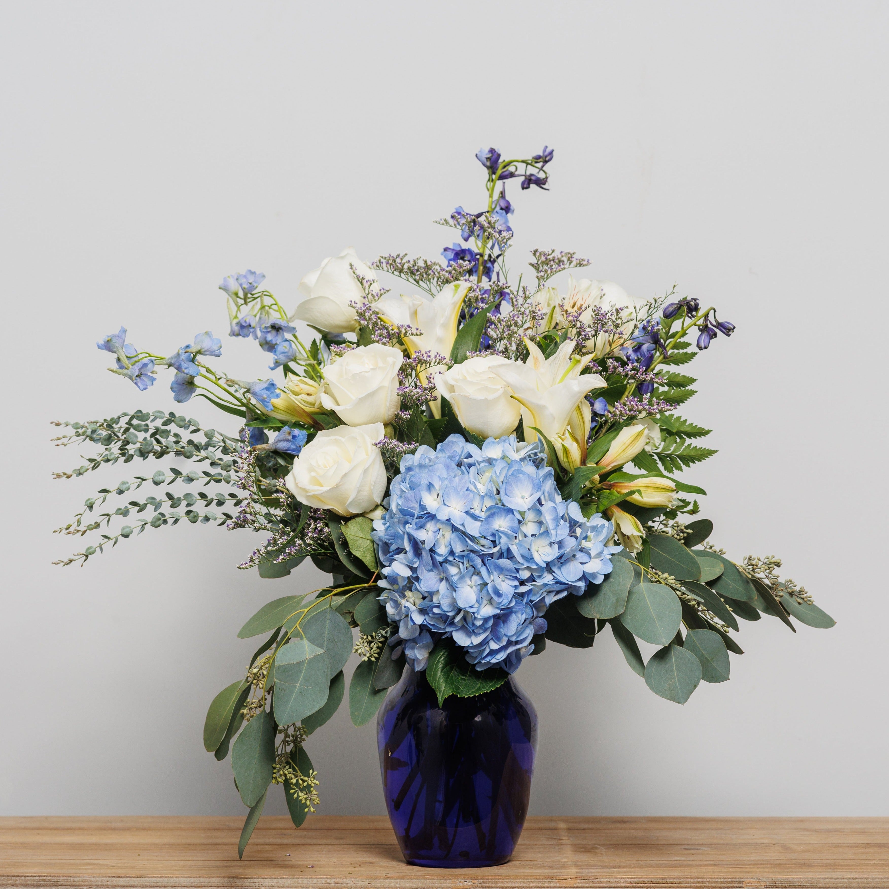An arrangement with white roses, white lilies and blue hydrangea in a blue vase.