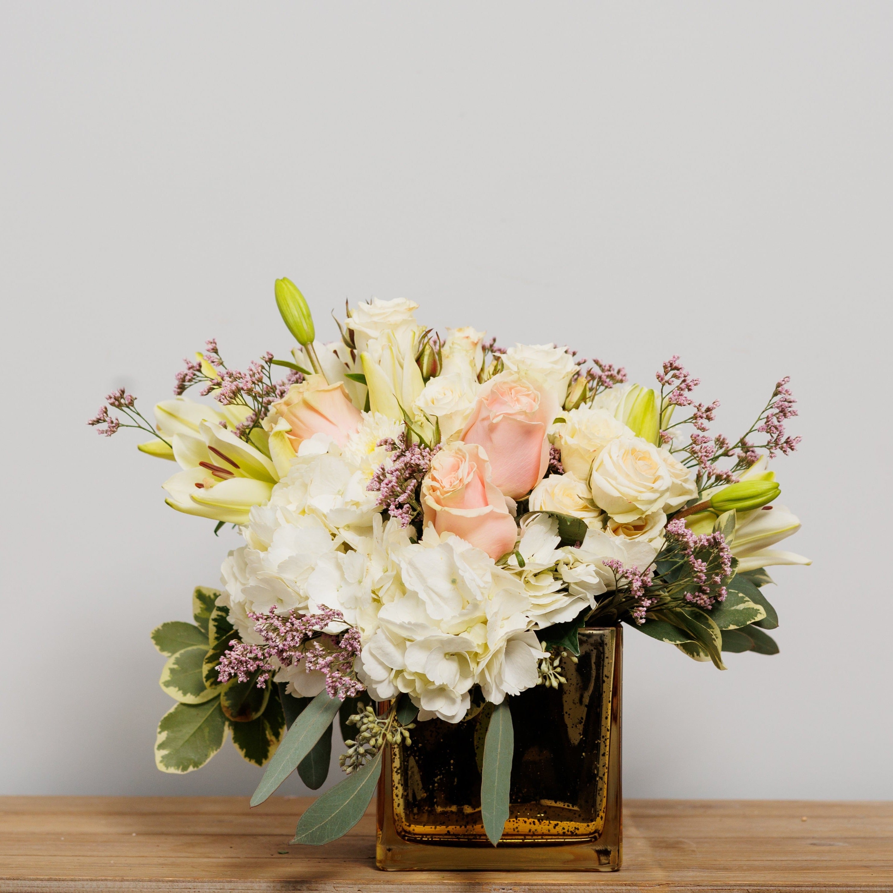 A flower arrangement with ivory and blush flowers in a golden cube.
