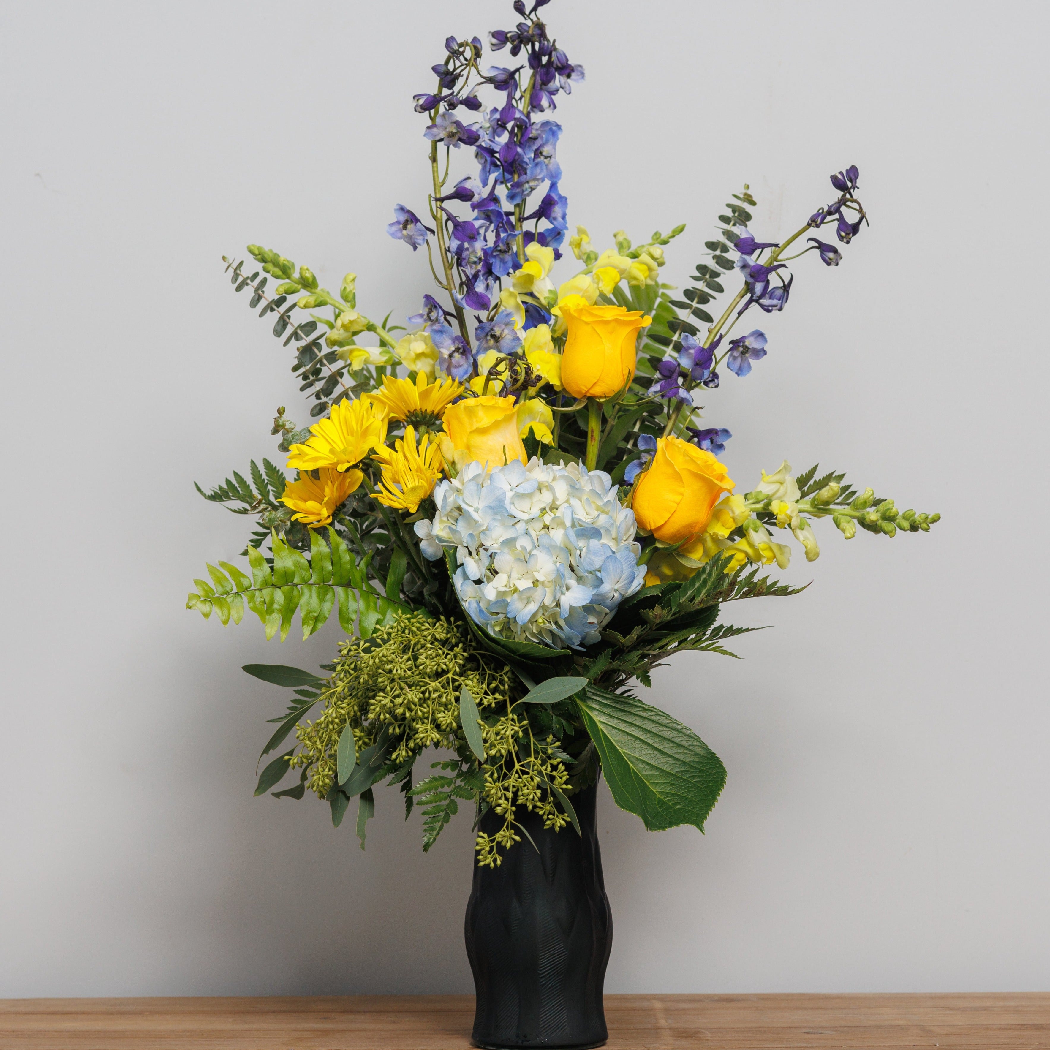 A vase arrangement with yellow roses, blue delphinium and blue hydrangea in a blue vase.