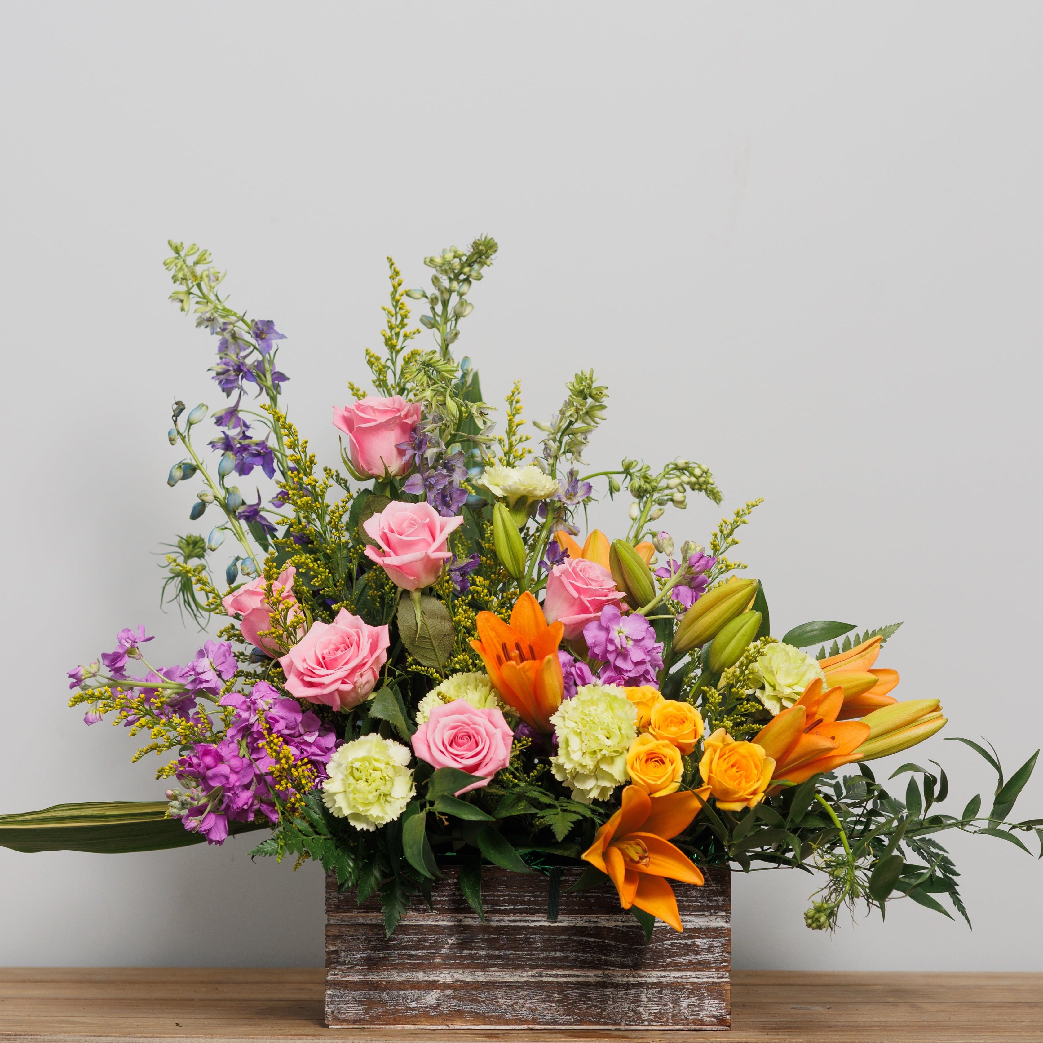 An arrangement with orange lilies, pink lilies and purple stock in a wooden box.