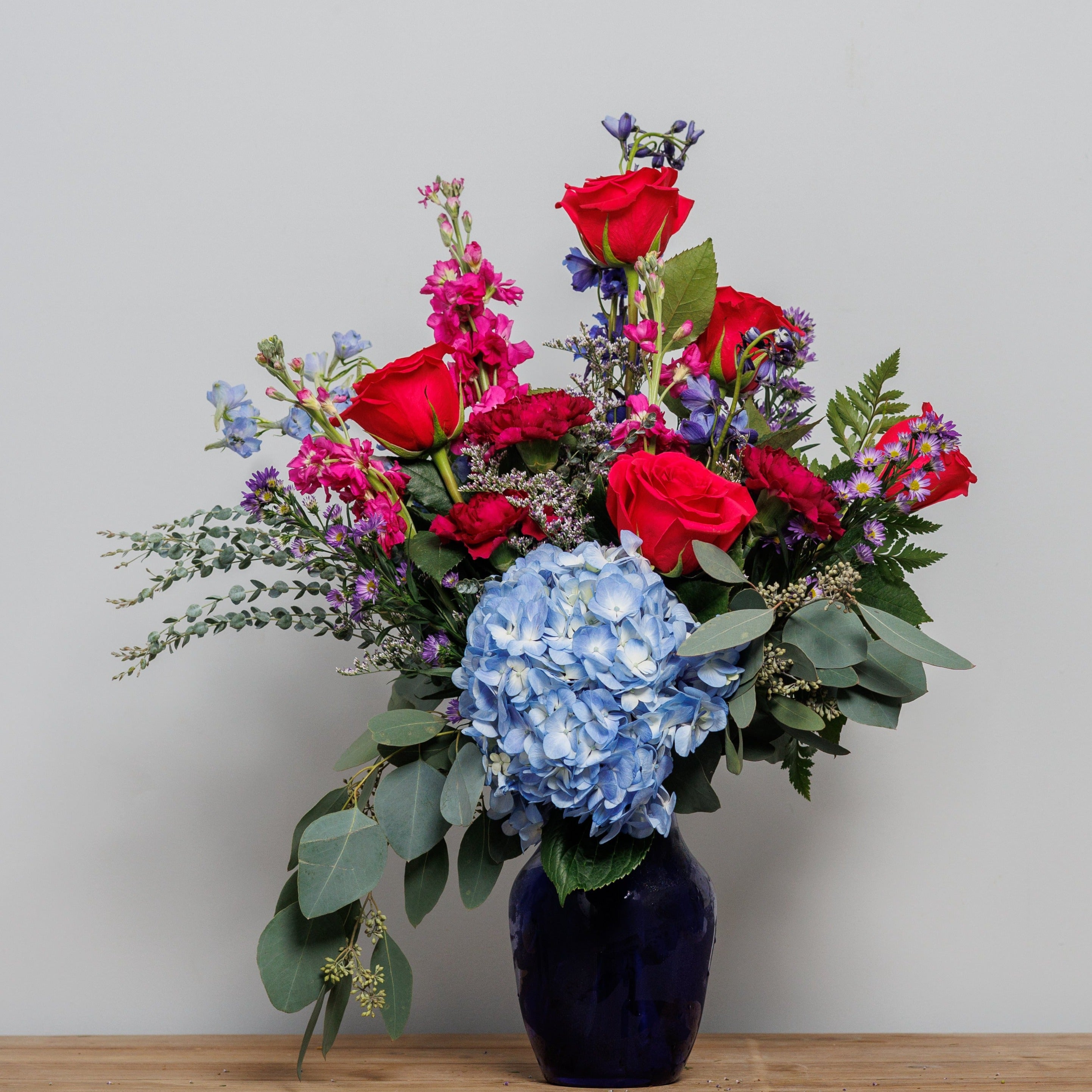 Hot pink roses with blue delphinium and a blue hydrangea in a blue vase.