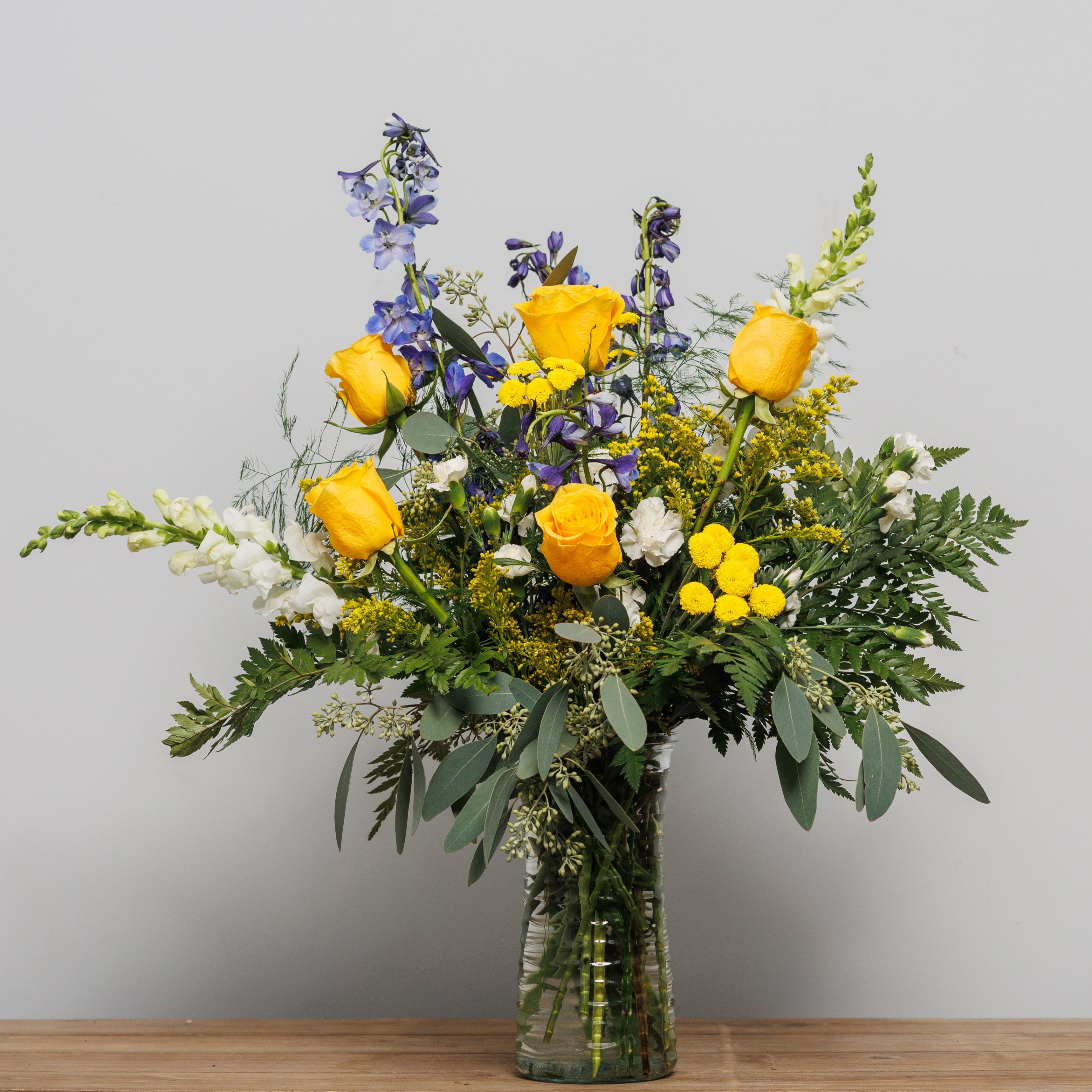 Yellow roses, blue delphinium and white snap dragons in a vase arrangement