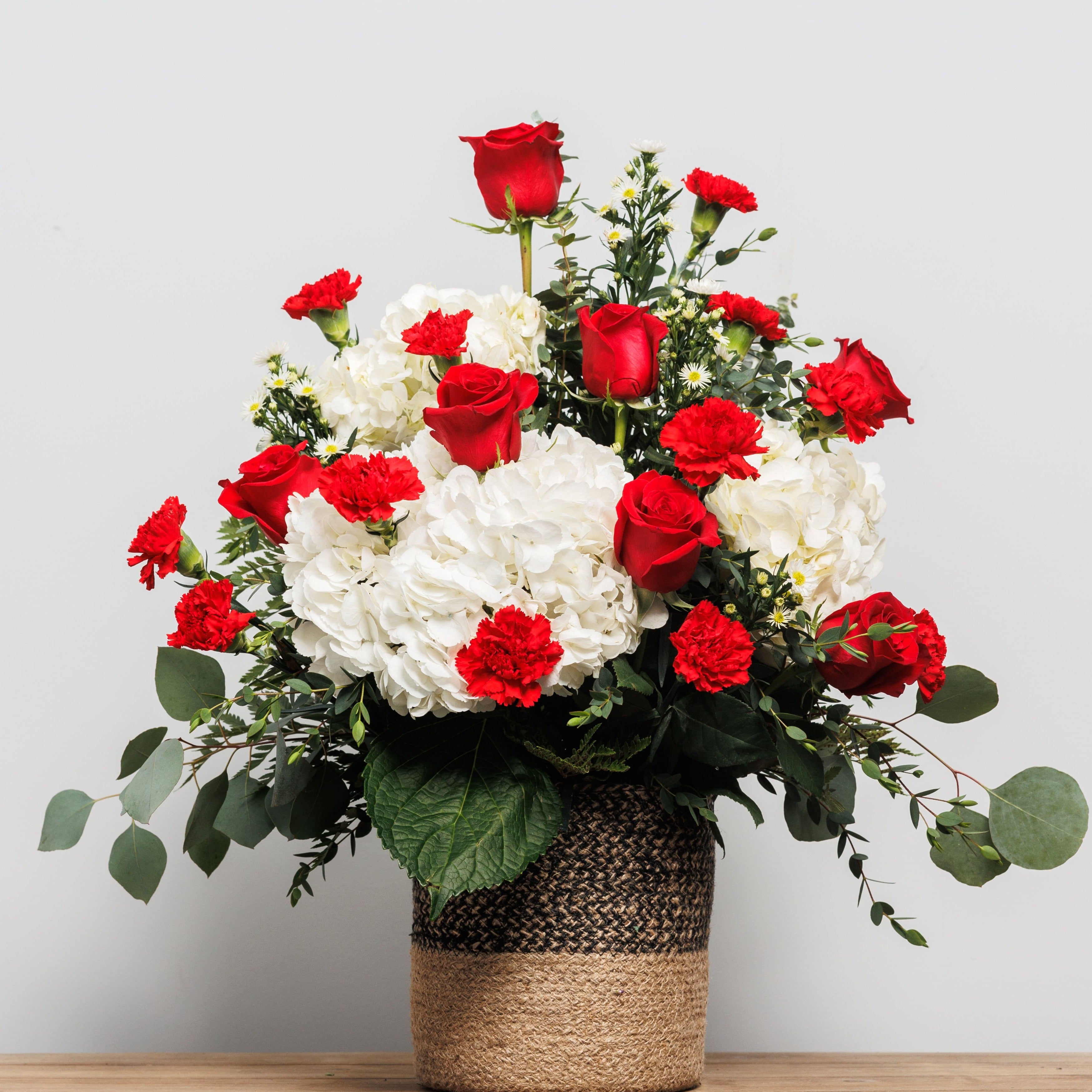 A basket arrangement with red roses, red carnations and white hydrangeas.