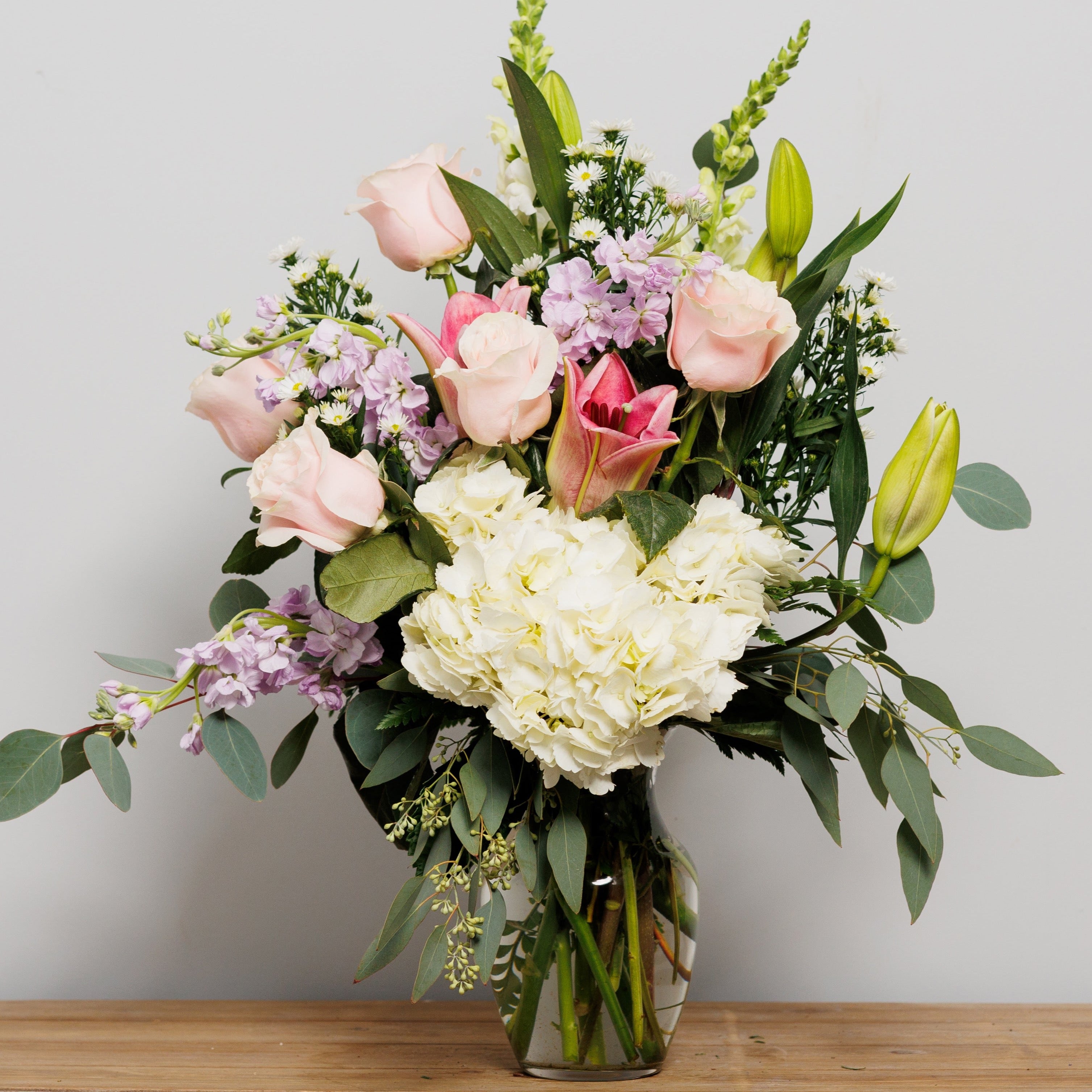 A vase arrangement with white hydrangea, pink roses and lilies, and lavender stock.