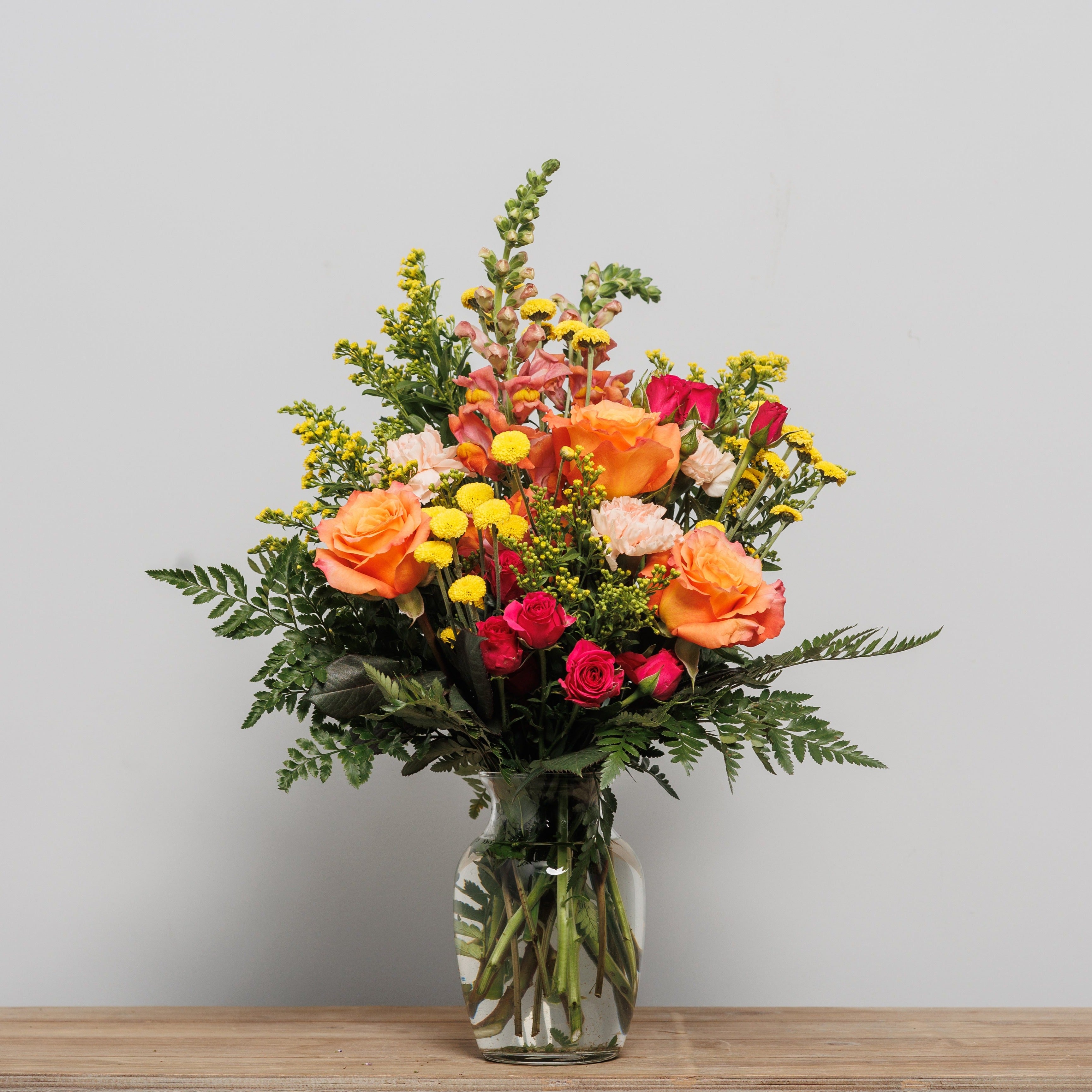 A bright flower arrangement with orange roses, yellow mums and pink snapdragons.